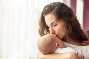 Young white mother holds a newborn in her arms and kisses the child feeling connected after postpartum anxiety and depression counseling in Roswell, GA