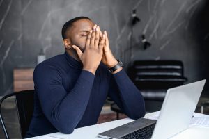 A man sits with hands over his face in front of a laptop. A therapist in Roswell, GA can offer support in finding a job with life transitions therapy in Roswell, GA. Learn more about individual counseling in Roswell, GA and more by contacting a young adult therapist in Roswell, GA today! 30076